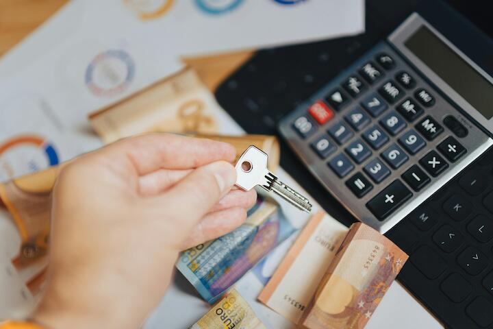 Eine Hand hält einen Schlüssel in der Hand. Unter der Hand ist ein Tisch mit Taschenrechner, Geld und vielen Zetteln