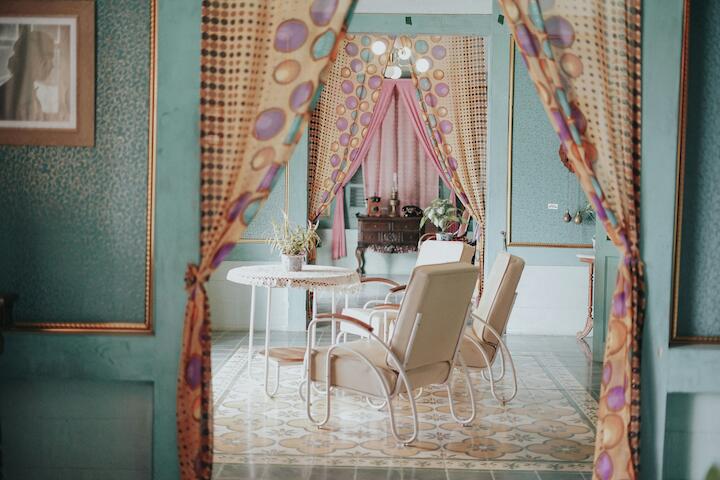 Eine farbenfrohes Zimmer mit bunten Tapeten im Rokoko-Stil, bunten Vorhängen und einem kleinen Tisch mit cremefarbenen Stühlen. Dahinter steht eine alte barocke Kommode mit Verzierungen in nussbraun..