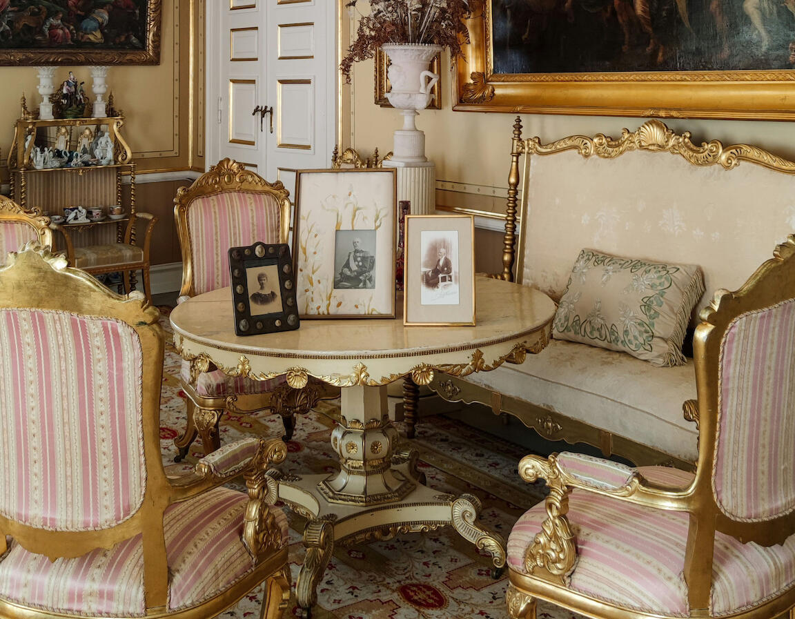 Ein Wohnzimmer mit barocken Elementen, wie Stühlen, einem runden Tisch, einem goldenen Bilderrahmen und ein Kronleuchter.