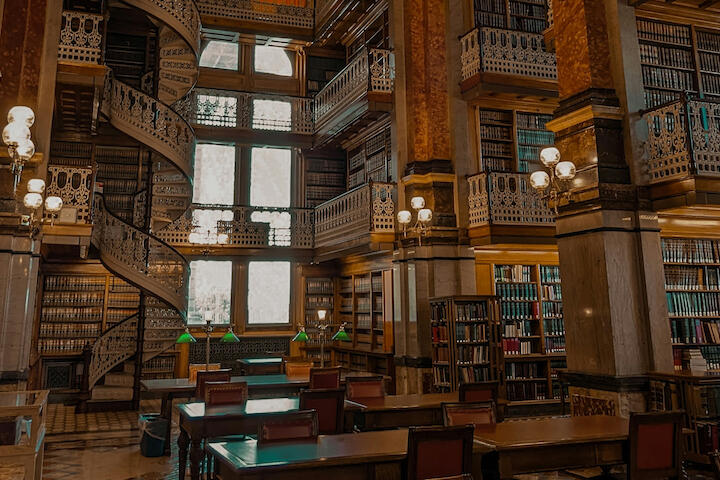 Eine Bibliothek mit hölzernen, verzierten Geländern, Büchern bis unter die Decke, schweren Tischen und ledernen Stühlen.