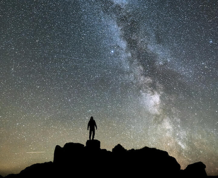Eine Person steht auf dunklen Felsen und brstaunt den Sternehimmel.