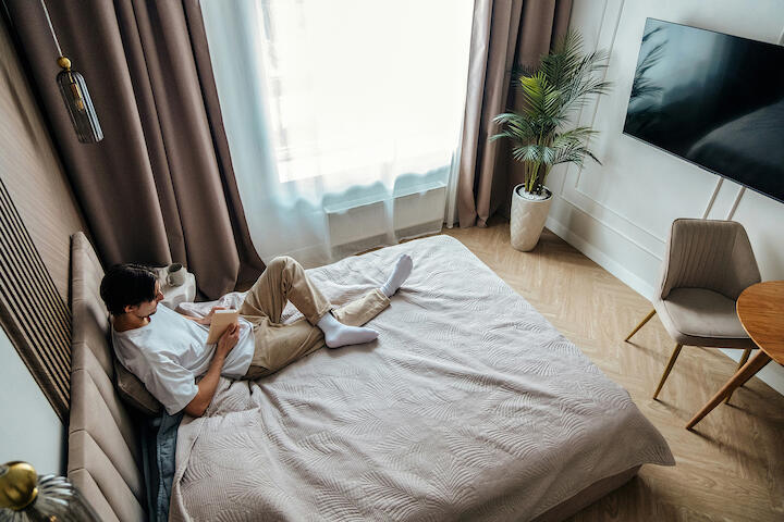 Ein Mann sitzt auf einem Bett und liest. In dem kleinen Zimmer sieht man noch den Fernseher an der Wand und einen Stuhl und ein Stück Tisch.
