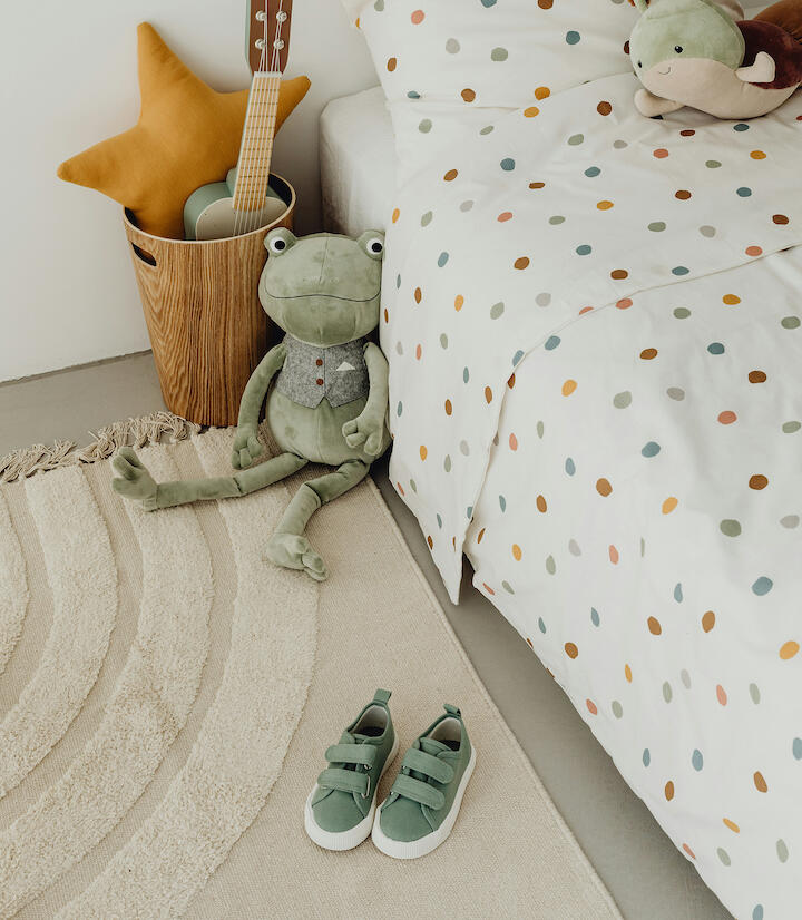 Man sieht einen Teppich vor einem Kinderbett. Darauf stehen grüne Stoffschuhe.